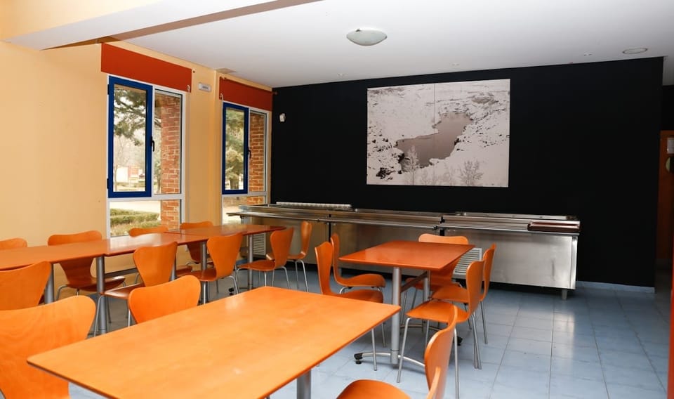 Comedor con mesas de madera clara y sillas naranjas frente a una pared oscura con un mapa