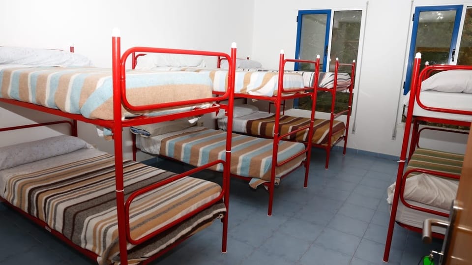 Habitación con tres literas metálicas rojas, colchas de rayas y ventana con contraventana azul