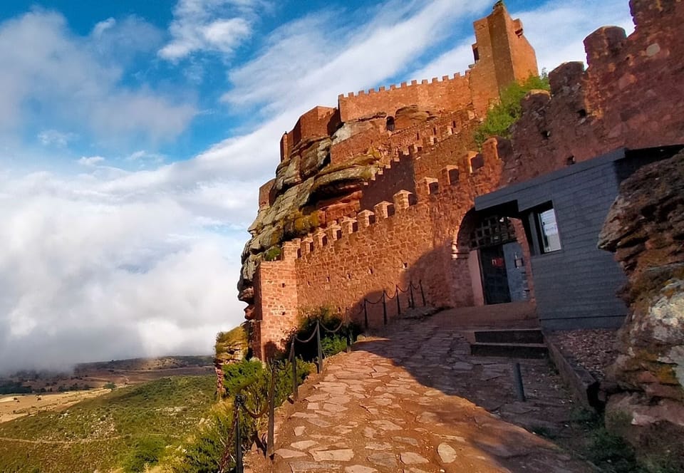 Muralla y torreón de piedra roja de Ojos Negros sobre el crestón rocoso, bajo un cielo con nubes