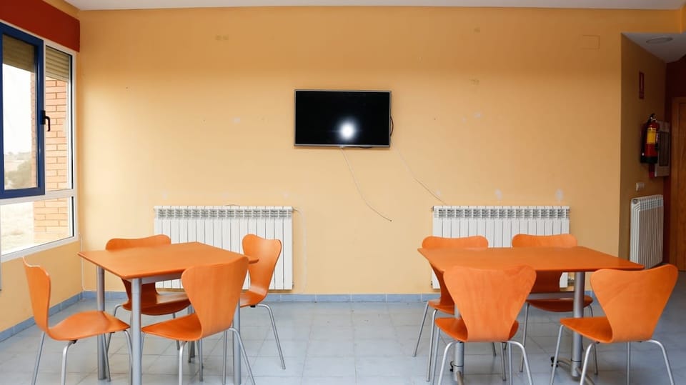 Sala polivalente con televisión, mesas y sillas naranjas y ventanas a ambos lados