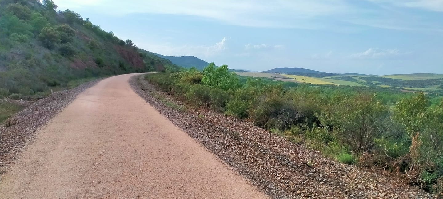 La Vía Verde de Ojos Negros trazando una curva de tierra rojiza entre matorral, con campos y sierra al fondo
